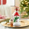 Glass bottle snowman ornament with red hat, green scarf, and faux snow for Christmas decor.