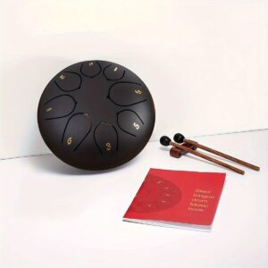 Black steel tongue drum with gold numbers, mallets, and red music book for meditation and relaxation.