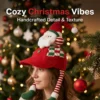 Red knitted Santa hat with plush Santa, striped ear flaps, and green/white checkerboard detail for festive wear.