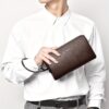 Men's brown pebbled leather clutch wallet with wrist strap, held by hand, for formal or casual use.