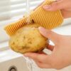 Yellow silicone vegetable scrubber with flexible bristles, used to clean a potato under running water.